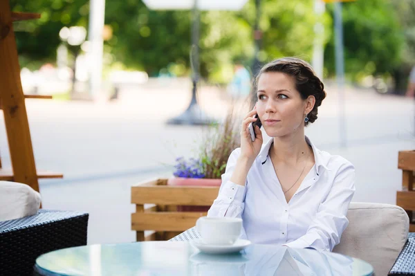 Portrait of young business woman sitting relaxed at outdoor cafe drinking coffee and talking using her cell phone