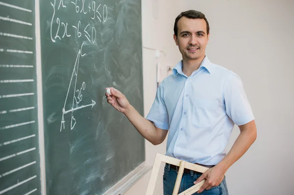 Young teacher or student holding triangle pointing at chalkboard with formula, looking to camera and smiling