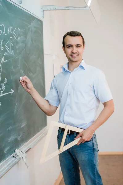 Young teacher or student holding triangle pointing at chalkboard with formula, looking to camera and smiling