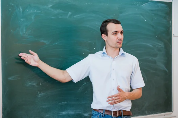 Portrait of young handsome student or teacher in a class pointing at blank chalkboard, talking and smiling