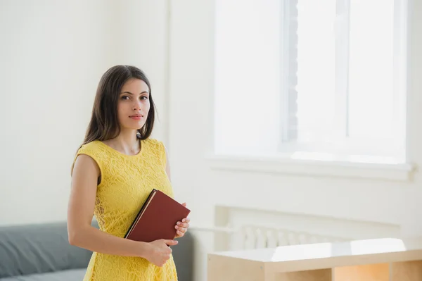 Happy pretty young female student with long blond hair clutching school books to her chest as she stands in front of the windows at classroom