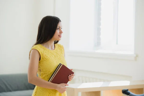 Happy pretty young female student with long black hair clutching school books to her chest as she stands in front of the windows at classroom