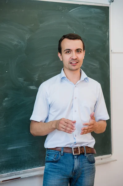 Portrait of young handsome student or teacher in a class pointing at blank chalkboard, talking and smiling