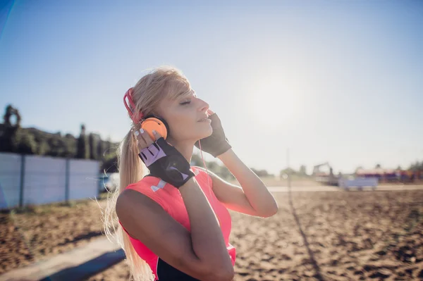 Sport outdoor photo of beautiful young blonde woman in pink colorful sport suit listening to music on headphones by the beach