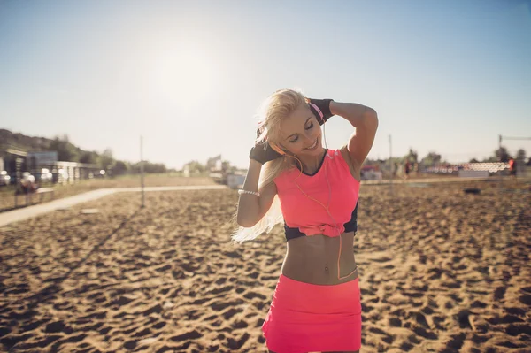 Sport outdoor photo of beautiful young blonde woman in pink colorful sport suit listening to music on headphones by the beach