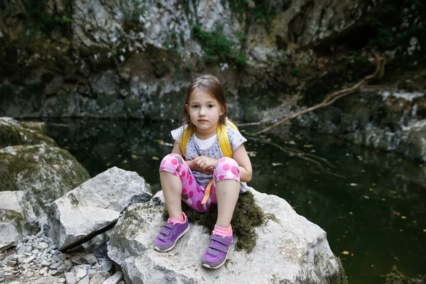 Happy little girl resting on a big rock by a small pond