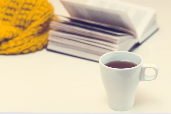 White cup of tea, book and scarf on light wooden desk.