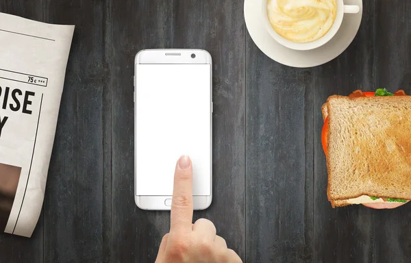 Smart phone with isolated display for mockup. Hand touching display. Newspaper, coffee and sandwich on table. Top view.