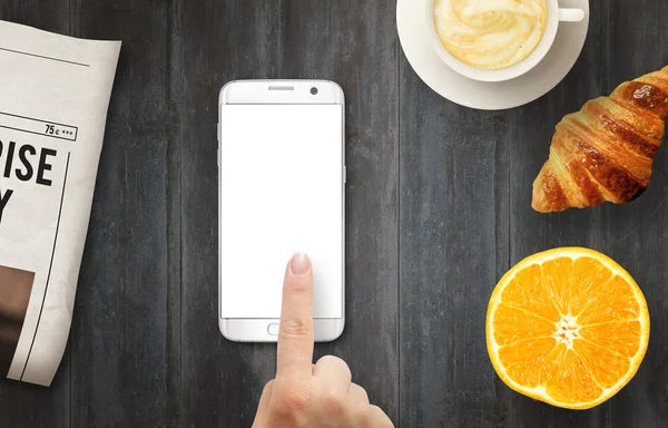 Smart phone with isolated display for mockup. Hand touching display. Newspapers, coffee, croissant and orange on table. Top view.