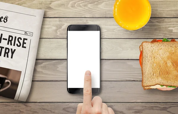 Smart phone with isolated display for mockup. Hand touching display. Newspaper, juice and sandwich on table. Top view.