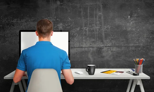 Man working on computer with isolated screen in office interior. Work desk with keyboard, mouse, cup of coffee, paper, pencils. Free space on wall for text.