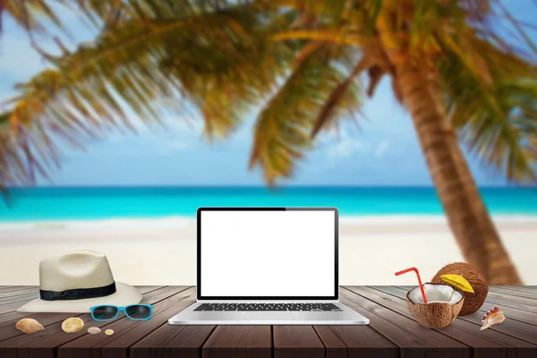 Isolated display of laptop on wooden table for mockup. Coconut, hat, shells, stones, sunglasses on table. Beach, sea, palm and blue sky in background.