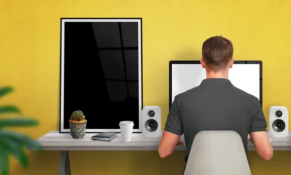 Man work on computer. Poster frame beside. Coffee, cactus, speakers on table. Free space on wall for text. Yellow wall in background.