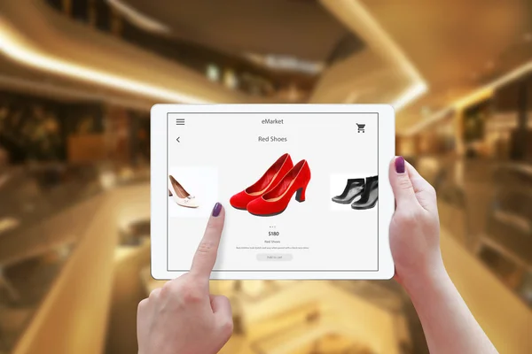 Woman online shopping with tablet. Holding device and choose red shoes. Shopping center in the background.