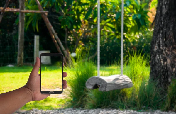 The hand of man hold mobile phone over blurred log swing hanging under the tree in garden