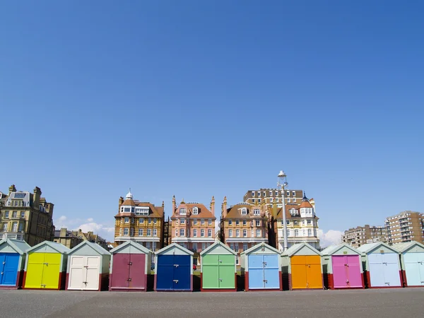 Brightly colored beach huts lined up along the beach with traditional architecture behind, under clear blue sky.