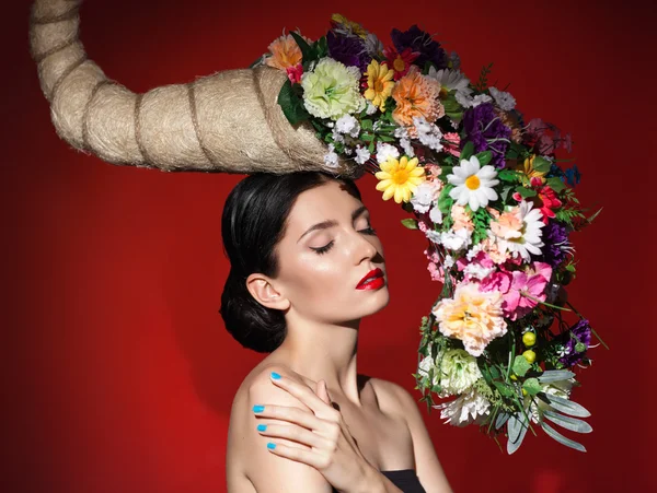 White woman posing in studio with closed eyes. Perfect makeup black eyeliner and red lipstick.Model is in incredible big hat with colorful flowers. Blue nails, red background.