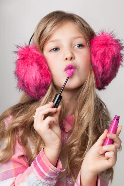 Beautiful little girl with long blonde hair wearing pink fur headphones, taking a pink lipstick on her hands.