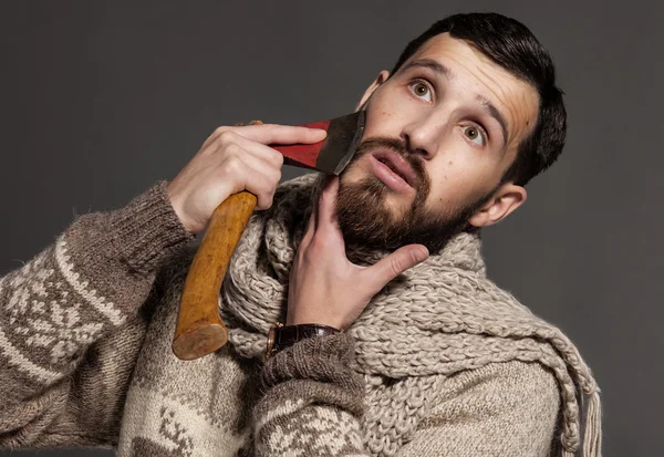 Making his beard perfect. Confident young bearded man shaving with ax and looking at camera while standing against grey background