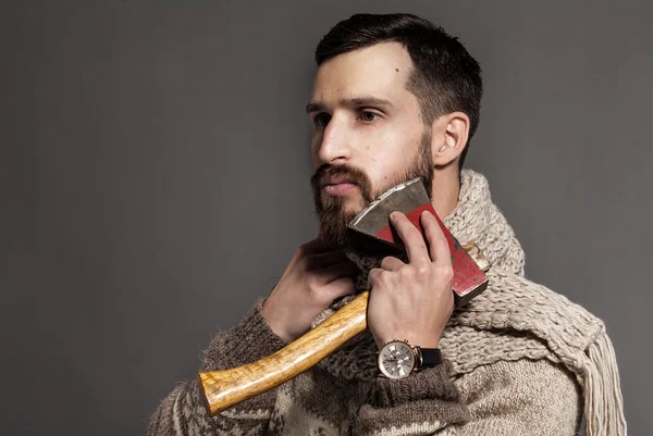 Making his beard perfect. Confident young bearded man shaving with ax and looking at camera while standing against grey background
