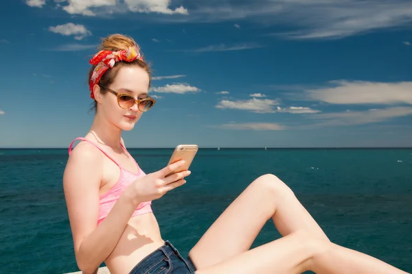 Attractive girl pin up model in pink top and jeans shorts with leopard sunglasses sunbathing and chating on smartphone