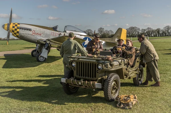 Ground crew sit in a WW2 jeep with a P-51 Mustang in the backgro