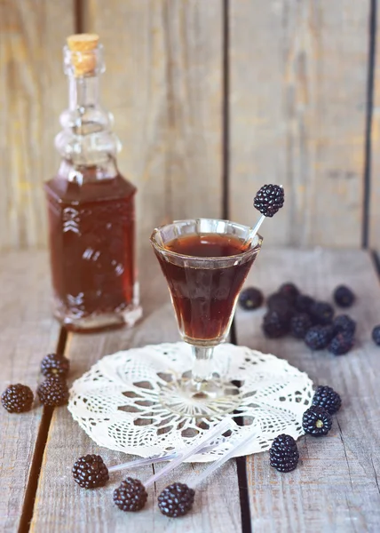 Blackberry liquor in the little glass and vintage bottle on the vintage napkin on the old wooden background, toned picture, selective picture