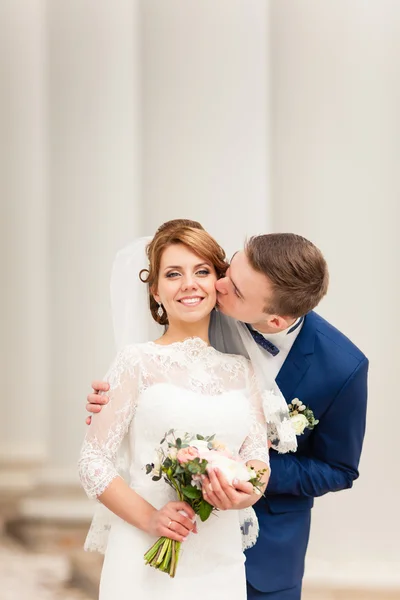 The groom kisses the bride that stands between the columns and holding a bridal bouquet