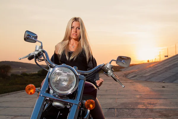 Young sexy blonde sitting on her motorcycle on the background of a summer sunset