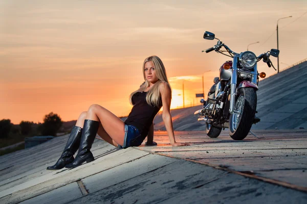 Young sexy blonde sitting near her motorcycle on the background of a summer sunset