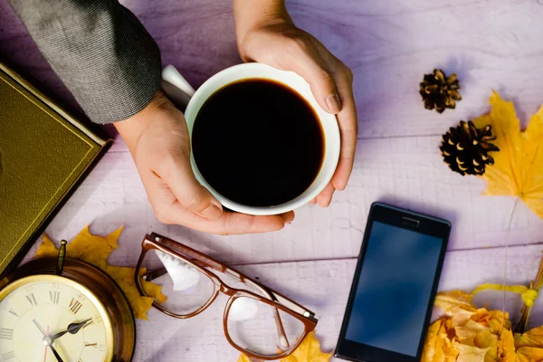 View from top on hands holding cup of coffee with mobile phone, glasses and alarm clock over wooden table background, close up picture