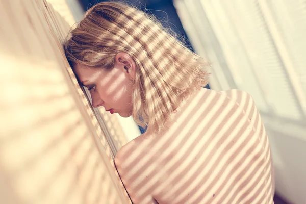 Enjoying sunshine: blond attractive topless girl with shadow on bare shoulders from sun light rays through shutters standing near wooden wall portrait picture