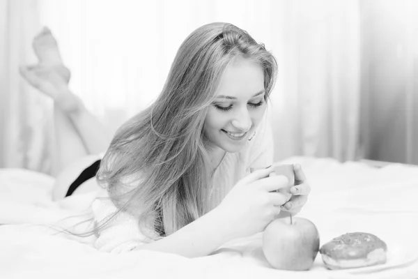 Black and white closeup portrait of beautiful sexy blonde young lady lying in bed smiling looking at plate with donut and apple on light window background