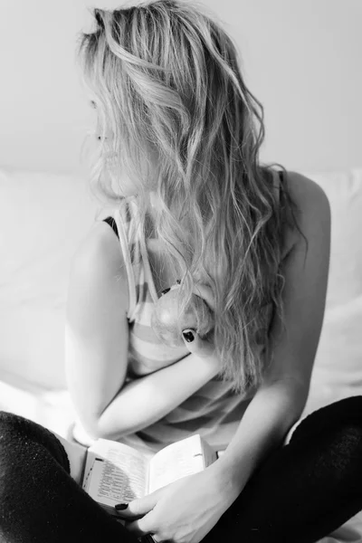 Portrait of sexy blonde girl having fun happy holding book and apple sitting on bed. Black and white image