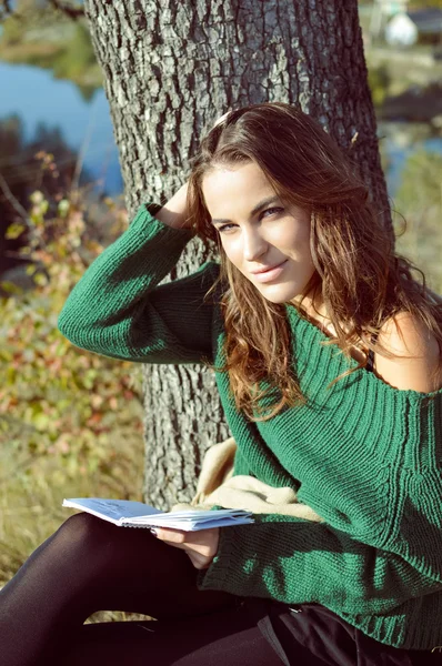 Young girl in sweater sitting under tree reading and smiling
