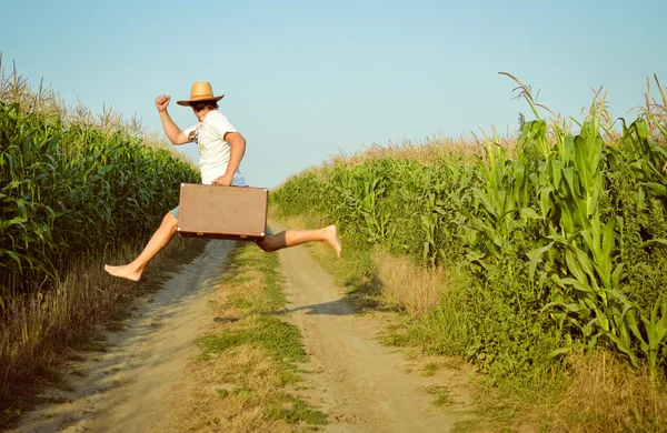 Rear view of man in straw hat enjoying his life jumping with suitcase on rural road.