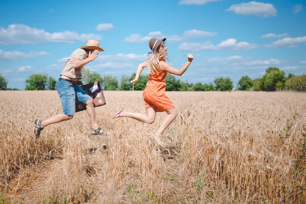 Happy romantic couple running with old suitcase in summer wheat field