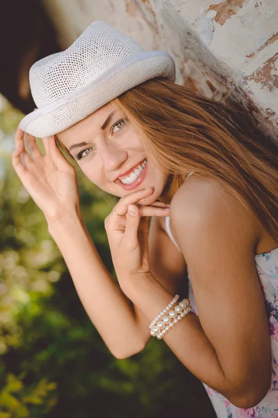 Beautiful young lady in white fedora hat smiling beside wall