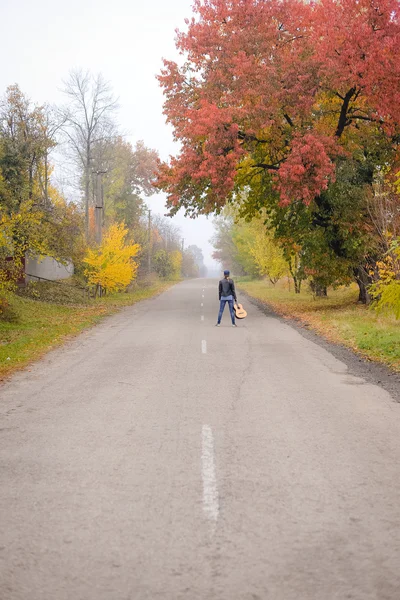 Beautiful young woman with guitar standing on autumn countryside road