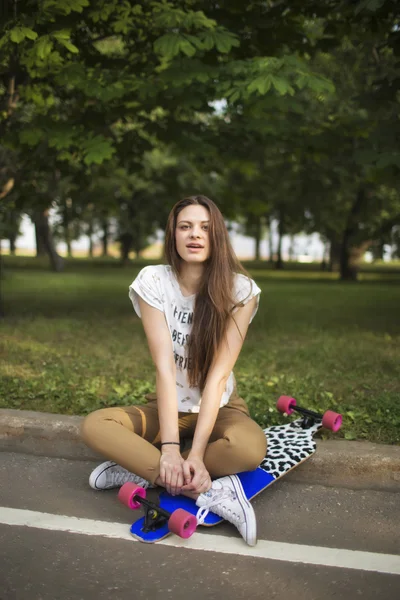 Young girl sitting cross-legged in the park next to the longboard and looking at the camera. Lifestyle outdoor