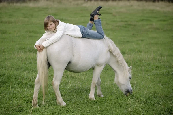 Little young girl in a white sweater and jeans lying on the back of a white pony. Lifestyle portrait