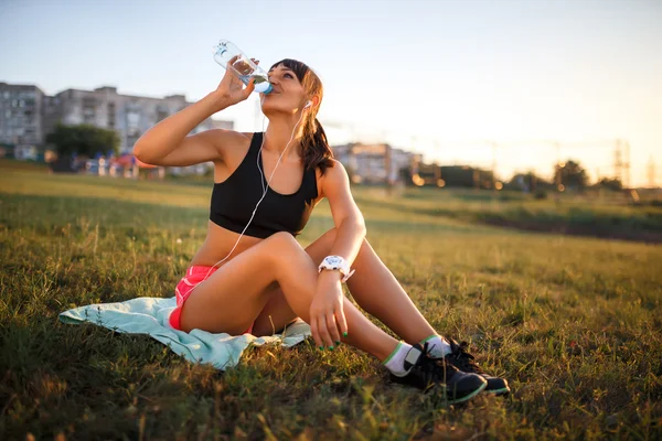 Sporty young woman drinking water. girl sits on a mat and listening music on headphones