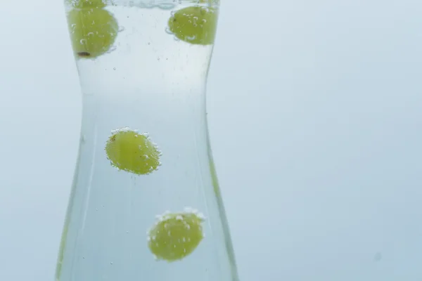 White wine grapes marbles in carafe with sparkling water surrounded by air bubbles, one in focus others in motion on blue neutral backdrop.