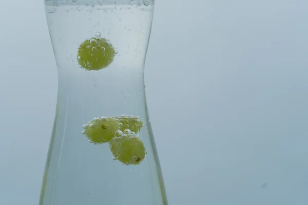 White wine grapes marbles in carafe with sparkling water surrounded by air bubbles in motion on blue neutral backdrop.