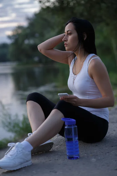 Woman runner listen music after workout and looking at sunset near pond