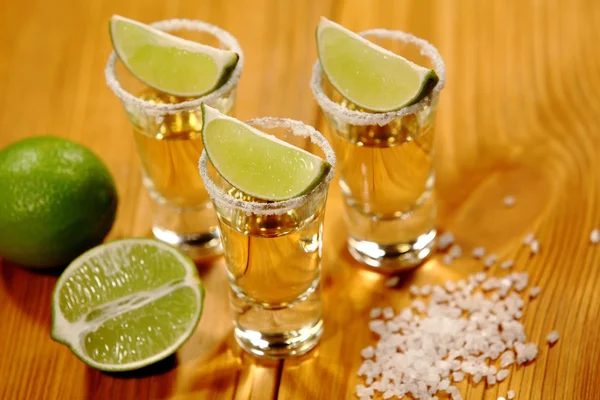 Three short glasses with alcohol next to a slice of lime and salt are on an old rustic table with vintage texture