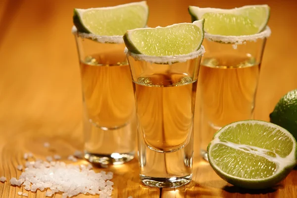 Three short glasses with alcohol next to a slice of lime and salt are on an old rustic table with vintage texture