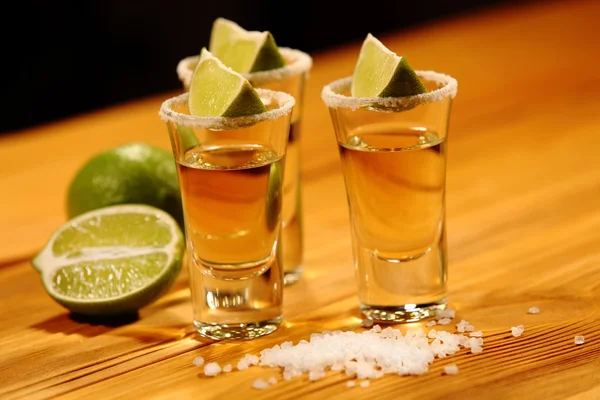 Three short glasses with alcohol next to a slice of lime and salt are on an old rustic table with vintage texture