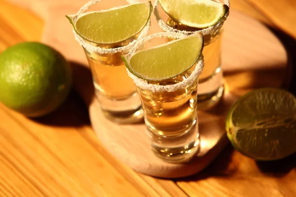 Three short glasses with alcohol next to a slice of lime and salt are on an old rustic table with vintage texture