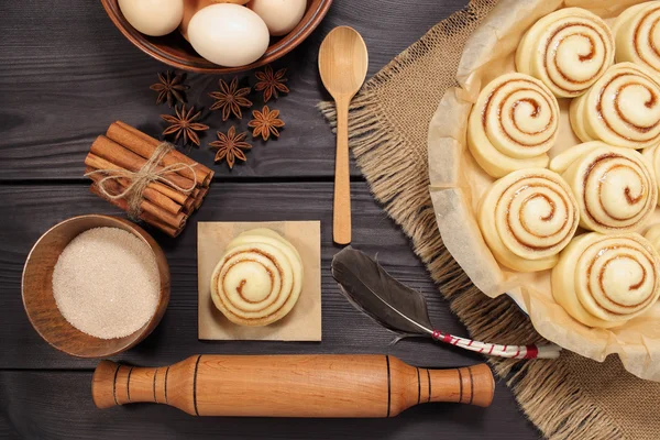 Raw cinnamon rolls prepared for roasting next to eggs and kitchen tools on a background of rustic table of black boards